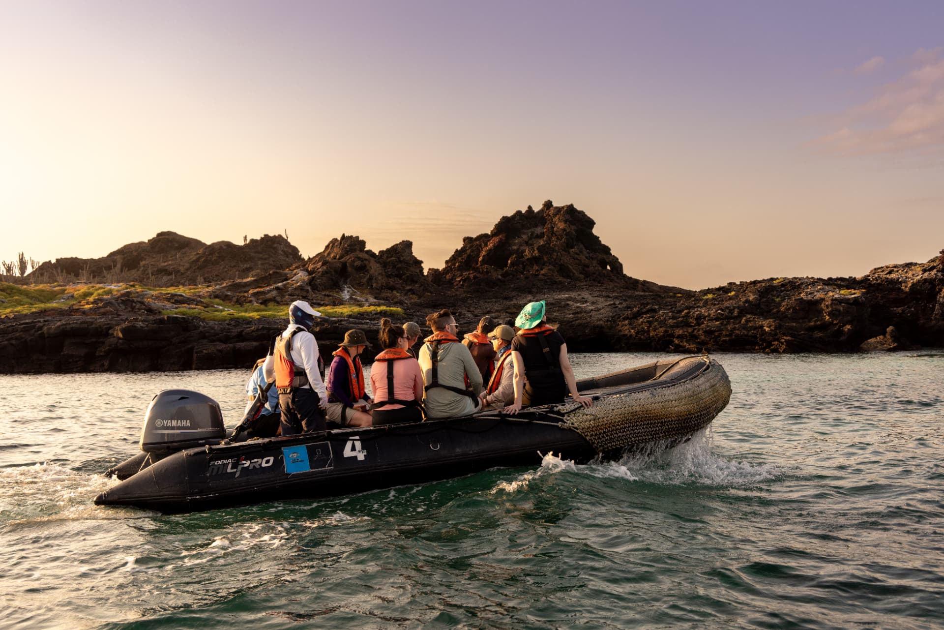 Naturalist-led exploration photography — guests aboard a Zodiac approaching a volcanic Galápagos shoreline at golden hour